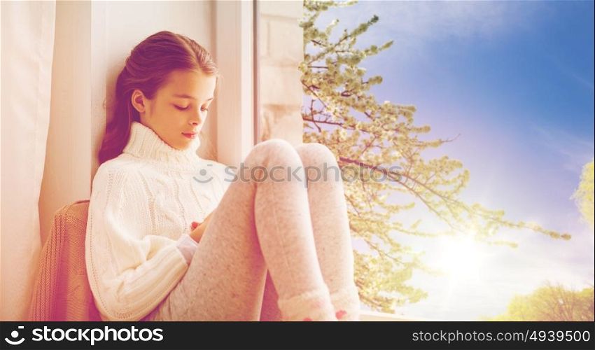 childhood, sadness and people concept - sad beautiful girl in sweater sitting at home window over natural spring cherry tree background. sad girl sitting on sill at home window