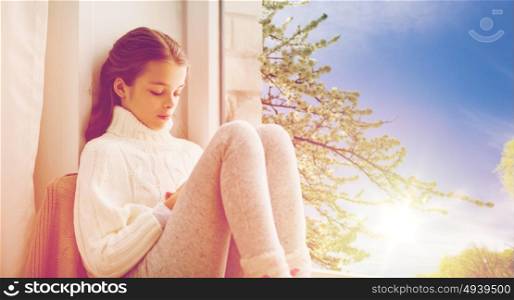 childhood, sadness and people concept - sad beautiful girl in sweater sitting at home window over natural spring cherry tree background. sad girl sitting on sill at home window