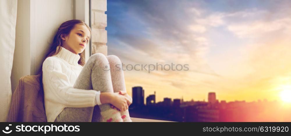 childhood, sadness and people concept - sad beautiful girl in sweater sitting on sill at home window over city background. sad girl sitting on sill at home window