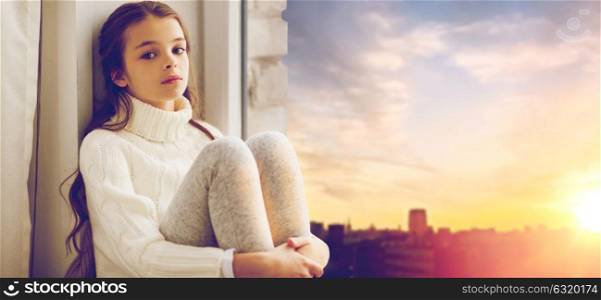 childhood, sadness and people concept - sad beautiful girl in sweater sitting on sill at home window over city background. sad girl sitting on sill at home window