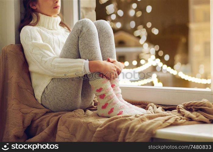 childhood, sadness and people concept - sad beautiful girl in sweater sitting on sill at home window over christmas lights background. sad girl sitting at home window on christmas