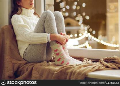 childhood, sadness and people concept - sad beautiful girl in sweater sitting on sill at home window over christmas lights background. sad girl sitting at home window on christmas
