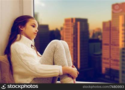 childhood, sadness and people concept - sad beautiful girl in sweater sitting on sill at home window over city skyscrapers background. sad girl sitting on sill at home window over city