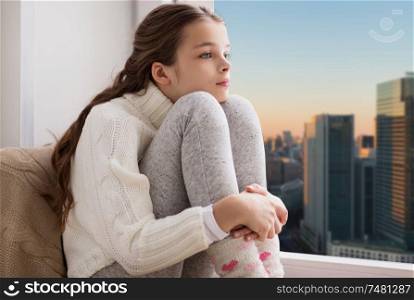 childhood, sadness and people concept - sad beautiful girl in sweater sitting on sill at home window over city skyscrapers background. sad girl sitting on sill at home window over city