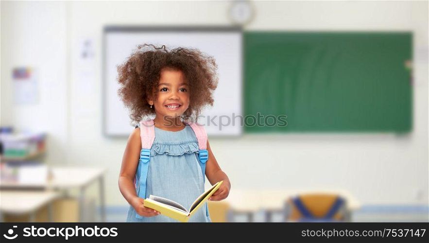childhood, school and education concept - happy little african american girl with book and backpack over classroom background. happy little african girl with book and backpack