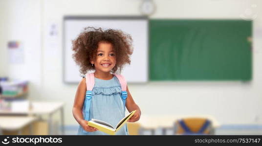 childhood, school and education concept - happy little african american girl with book and backpack over classroom background. happy little african girl with book and backpack