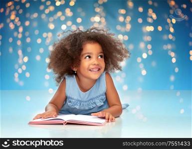 childhood, school and education concept - happy smiling little african american girl reading book over festive lights on blue background. smiling little african american girl reading book