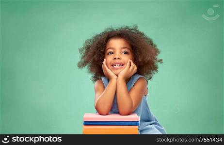 childhood, school and education concept - happy smiling little african american girl with pile of books over green chalk board background. little african american schoolgirl with books