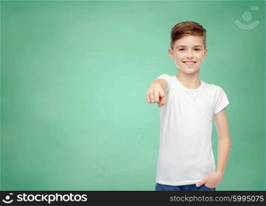 childhood, school, education, advertisement and people concept - happy student boy in white t-shirt and jeans pointing finger to you over green school chalk board background