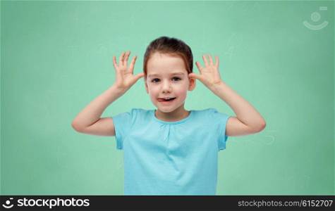 childhood, school, education, fun and people concept - happy little girl having fun and making ears over green chalk board background