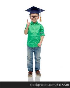 childhood, school, education, knowledge and people concept - happy boy in bachelor hat or mortarboard and eyeglasses pointing finger up