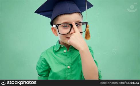 childhood, school, education, knowledge and people concept - happy boy in bachelor hat or mortarboard and eyeglasses over green school chalk board background