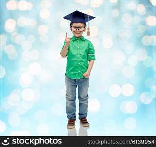 childhood, school, education, knowledge and people concept - happy boy in bachelor hat or mortarboard and eyeglasses pointing finger up over blue holidays lights background
