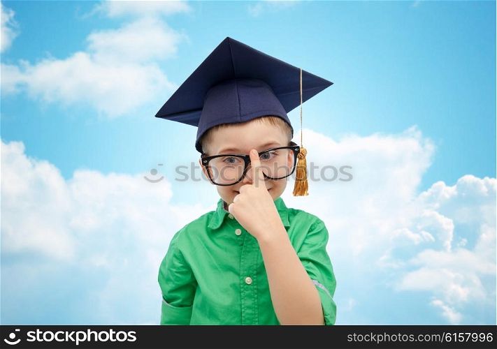 childhood, school, education, knowledge and people concept - happy boy in bachelor hat or mortarboard and eyeglasses over blue sky and clouds background