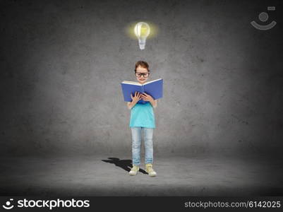 childhood, school, education, knowledge and people concept - happy little girl in eyeglasses reading book over concrete room background with light bulb symbol