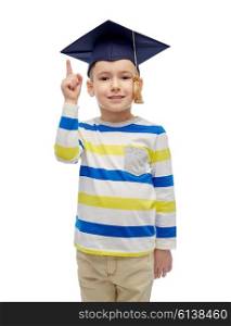 childhood, school, education, learning and people concept - happy boy in bachelor hat or mortarboard pointing finger up