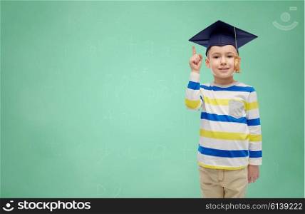 childhood, school, education, learning and people concept - happy boy in bachelor hat or mortarboard pointing finger up over green chalk board background