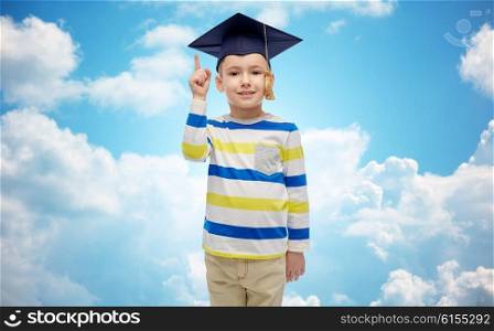 childhood, school, education, learning and people concept - happy boy in bachelor hat or mortarboard pointing finger up over blue sky and clouds background