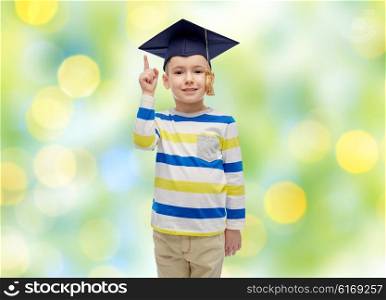 childhood, school, education, learning and people concept - happy boy in bachelor hat or mortarboard pointing finger up over summer green lights background