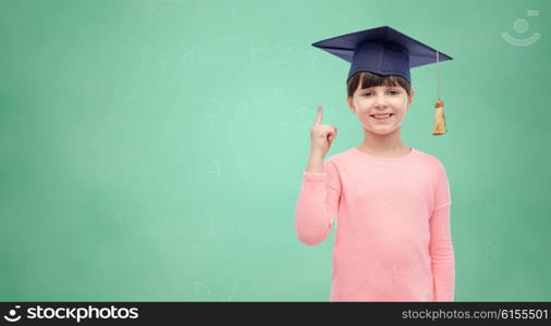 childhood, school, education, learning and people concept - happy girl with in bachelor hat or mortarboard over green school chalk board background