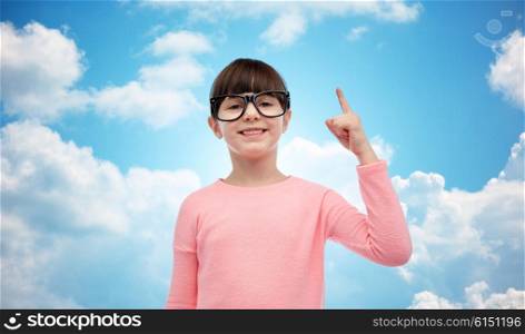 childhood, school, education, vision and people concept - happy little girl in eyeglasses pointing finger up over blue sky and clouds background