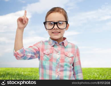 childhood, school, education, vision and people concept - happy little girl in eyeglasses pointing finger up over blue sky and green field background