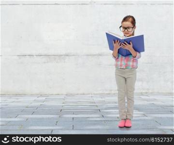 childhood, school, education, vision and people concept - happy little girl in eyeglasses reading book over urban concrete background
