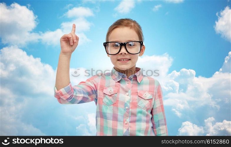 childhood, school, education, vision and people concept - happy little girl in eyeglasses pointing finger up over blue sky and clouds background