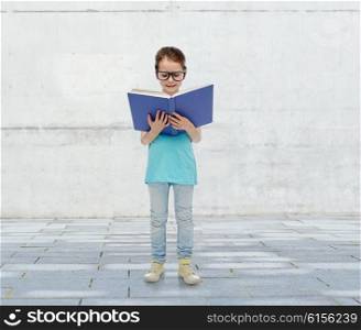 childhood, school, education, vision and people concept - happy little girl in eyeglasses reading book over urban concrete background