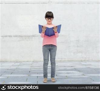 childhood, school, education, vision and people concept - happy little girl in eyeglasses reading book over urban city street background