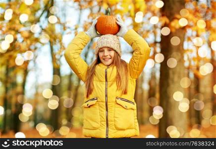 childhood, season and people concept - happy girl with pumpkin at autumn park over festive lights. happy girl with pumpkin at autumn park
