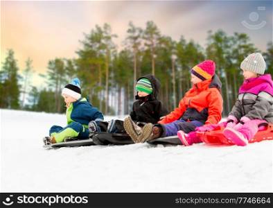 childhood, sledging and season concept - group of happy little children sliding on sleds in winter over snowy forest or park background. happy little children sliding on sleds in winter