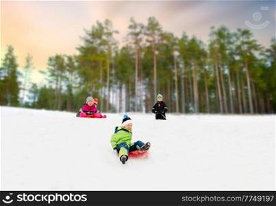 childhood, sledging and season concept - group of happy little children sliding on sleds down snow hill in winter over snowy forest or park background. kids sliding on sleds down snow hill in winter