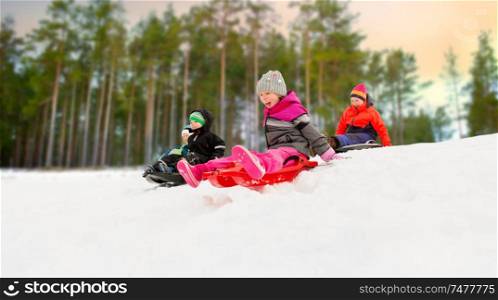 childhood, sledging and season concept - group of happy little kids sliding on sleds down snow hill in winter over forest background. kids sliding on sleds down snow hill in winter