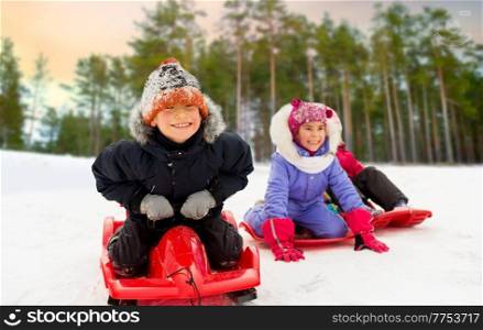 childhood, sledging and season concept - group of happy little kids sliding down on sleds in winter over snowy forest or park background. happy little kids sliding down on sleds in winter