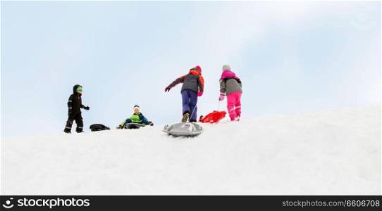 childhood, sledging and season concept - group of happy little kids with sleds climbing snow hill in winter. kids with sleds climbing snow hill in winter