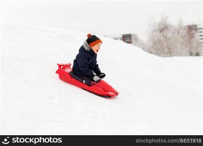 childhood, sledging and season concept - happy little boy sliding on sled down snow hill outdoors in winter. happy boy sliding on sled down snow hill in winter