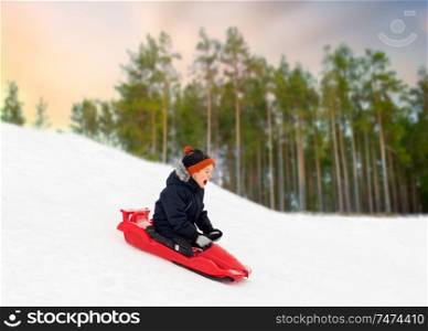 childhood, sledging and season concept - happy little boy sliding on sled down snow hill outdoors in winter over forest background. happy boy sliding on sled down snow hill in winter