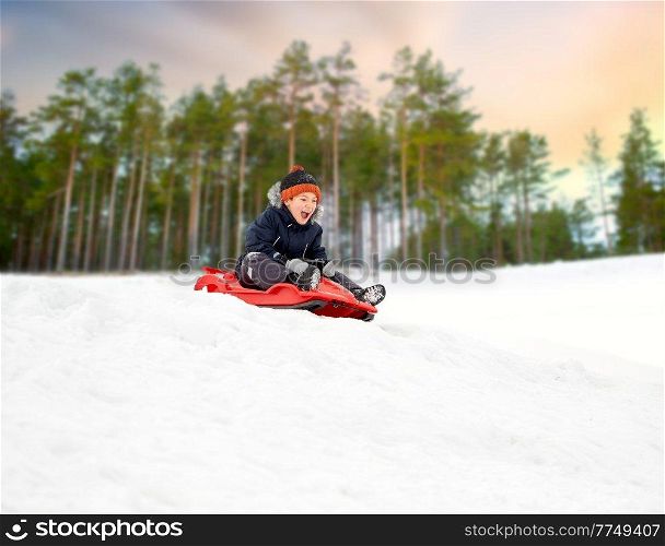 childhood, sledging and season concept - happy little boy sliding on sled down snow hill outdoors in winter over snowy park or forest background. happy boy sliding on sled down snow hill in winter