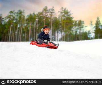 childhood, sledging and season concept - happy little boy sliding on sled down snow hill outdoors in winter over snowy park or forest background. happy boy sliding on sled down snow hill in winter