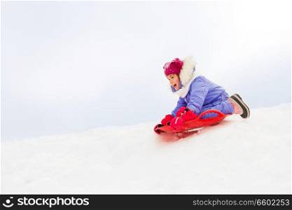 childhood, sledging and season concept - happy little girl sliding down on snow saucer sled outdoors in winter. girl sliding down on snow saucer sled in winter