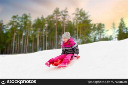 childhood, sledging and season concept - happy little girl sliding down on sled outdoors over winter forest background. happy little girl sliding down on sled in winter