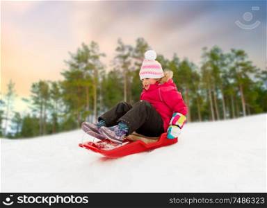 childhood, sledging and season concept - happy little girl sliding down on sled outdoors over winter forest background. happy little girl sliding down on sled in winter