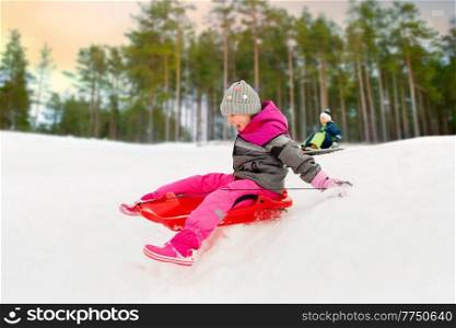 childhood, sledging and season concept - happy little girl sliding down on sled outdoors in winter over snowy park or forest background. happy little girl sliding down on sled in winter