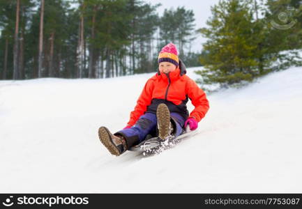 childhood, sledging and season concept - happy little girl sliding down on snow saucer sled outdoors in winter over snowy park or forest background. girl sliding down on snow saucer sled in winter