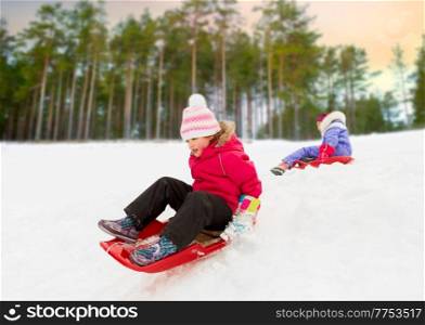 childhood, sledging and season concept - happy little girl sliding down on sled outdoors in winter over snowy park or forest background. happy little girl sliding down on sled in winter
