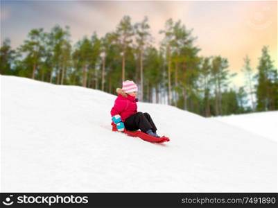 childhood, sledging and season concept - happy little girl sliding down snow hill on sled outdoors in winter over forest background. happy little girl sliding down on sled in winter