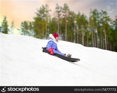 childhood, sledging and season concept - happy little girl sliding on sled down snow hill outdoors in winter over forest background. happy little girl sliding down on sled in winter