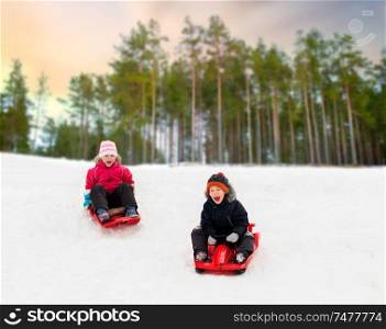 childhood, sledging and season concept - happy little kids sliding on sleds down snow hill in winter over forest background. happy kids sliding on sleds down hill in winter