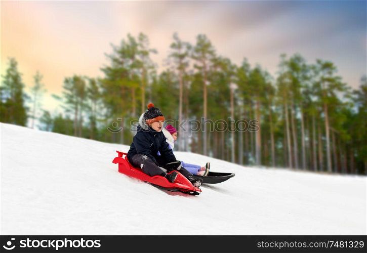 childhood, sledging and season concept - happy little kids sliding on sleds down snow hill over winter forest background. happy kids sliding on sleds down hill in winter